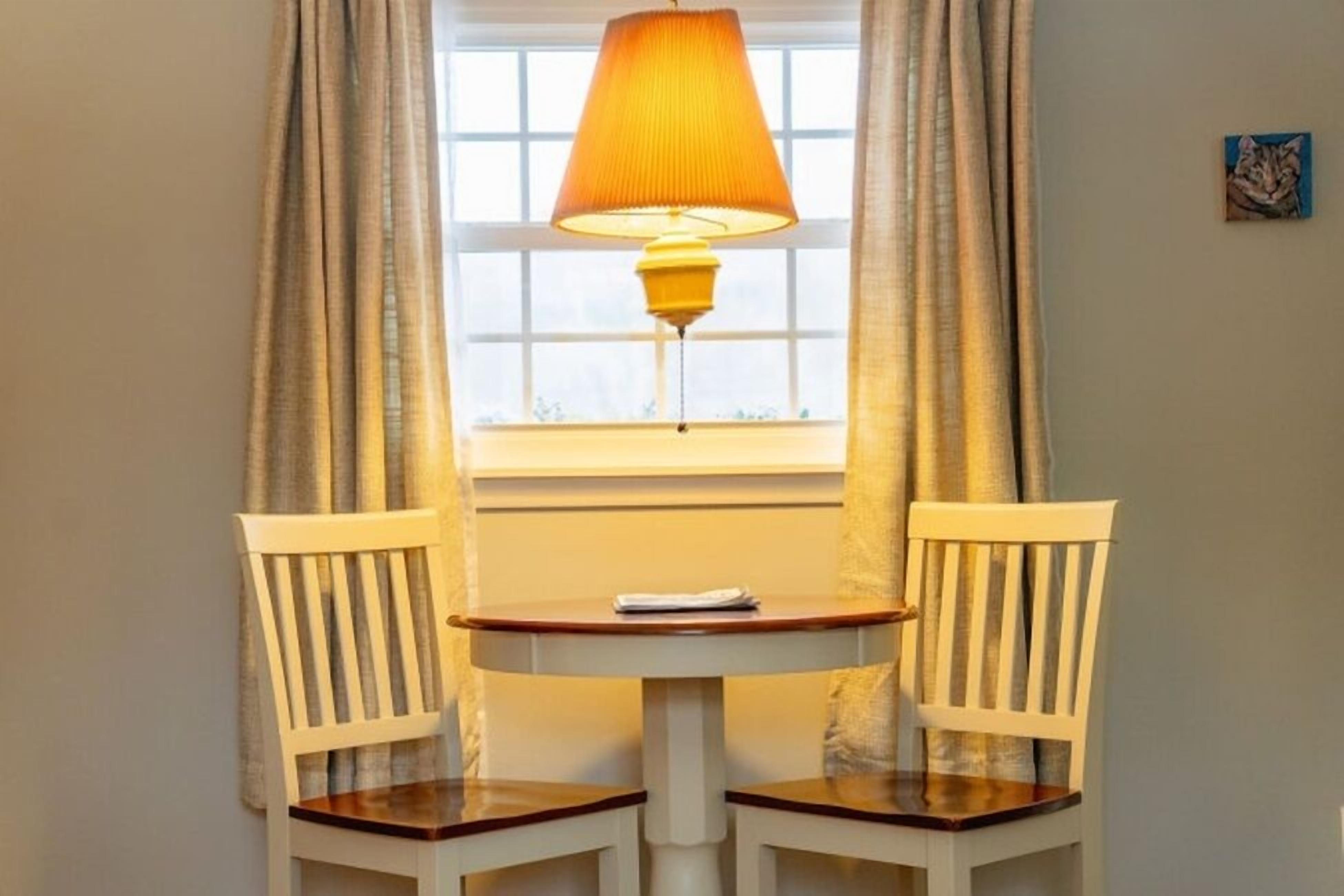 A room with a table and two chairs, a window with curtains, and a lamp.
