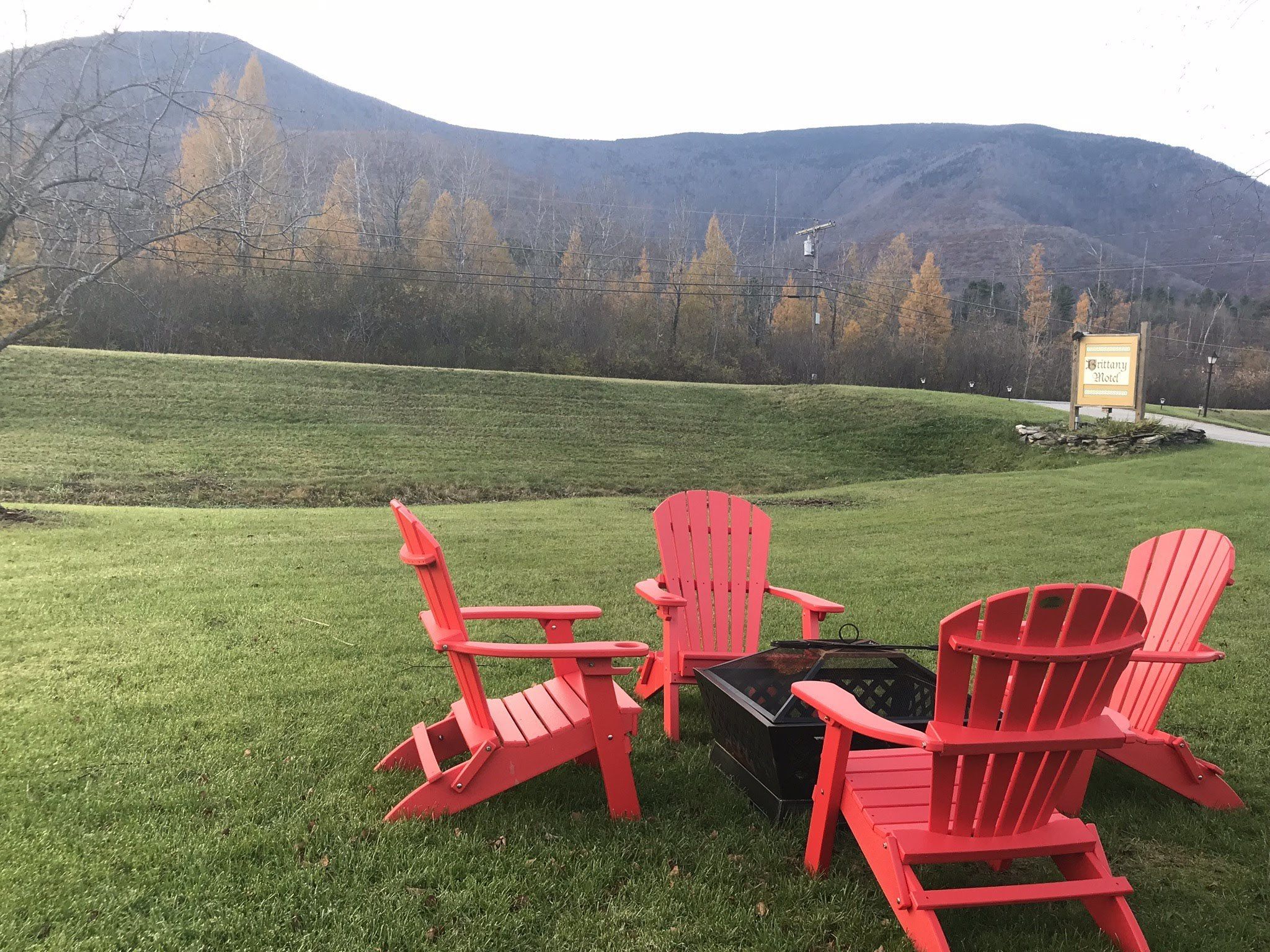 Un espacio cubierto de césped con tres sillas Adirondack rojas y un brasero negro. Al fondo, un cartel para el Motel Brittany.