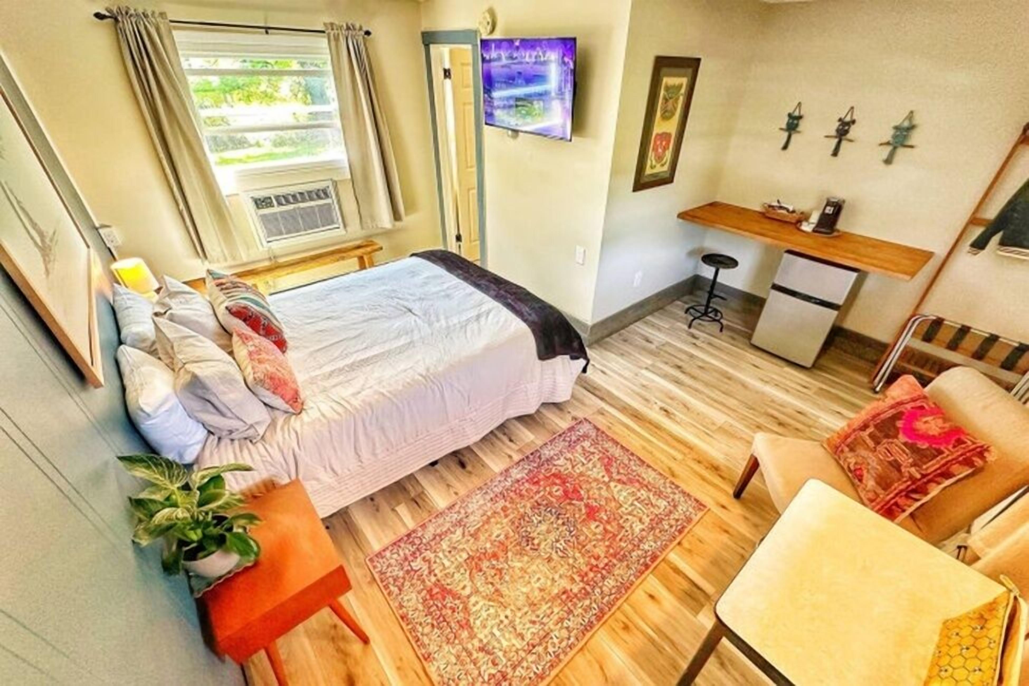 Ein kleines Hotelzimmer mit Bett, Schreibtisch, Stuhl, Tisch, Fernseher und einem Fenster mit Vorhängen.