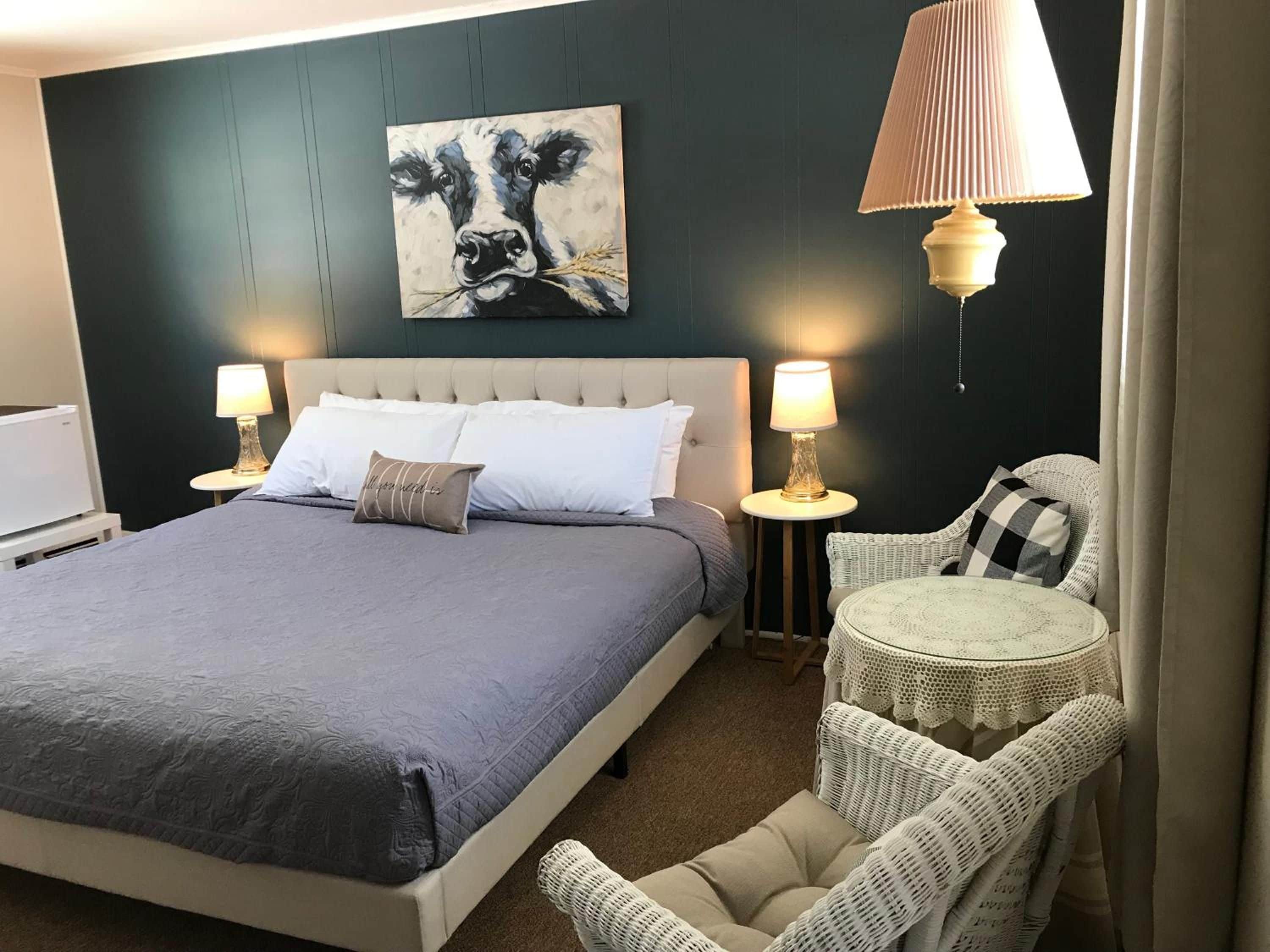 A hotel room with a large bed, two bedside lamps, a small round table, a wicker chair, and a painting on the wall.