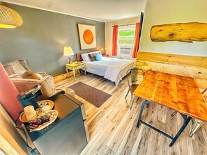 Una habitación de hotel acogedora con cama, una mesa de madera, una silla, una mesita con aperitivos, una lámpara y un amplio ventanal con cortinas.