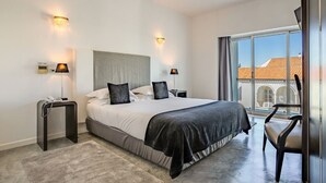 Habitación de hotel moderna con una cama grande, dos mesitas de noche, una silla y vistas de la ciudad a través de amplios ventanales.