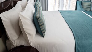 Un lit bien fait, avec une literie blanche et turquoise, un plaid turquoise, et une vue sur la plage par la fenêtre.