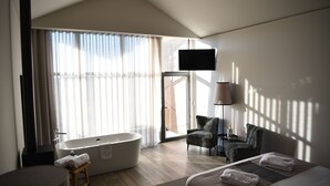Een moderne hotelkamer met een groot raam, een ligbad, een flatscreen televisie, een stoel en een bed met handdoeken.