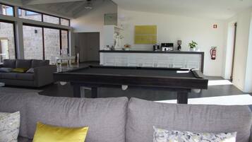 A modern living room with a pool table, grey sofa, and a kitchen area with white cabinets and a yellow accent wall.