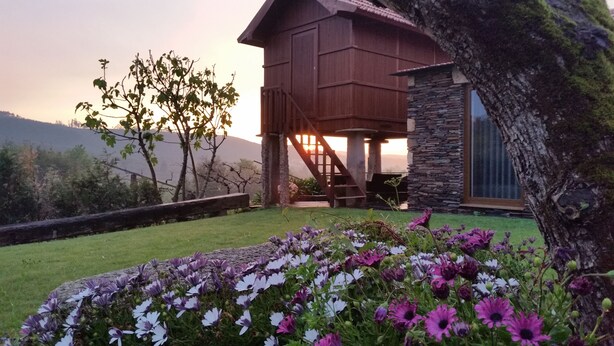 Een houten hut met een stenen fundering, omringd door een verzorgde tuin met paarse bloemen, met heuvels en een zonsondergang op de achtergrond.
