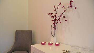 Una habitación con una mesa blanca, tres velas encendidas, un florero con una rama de flores rojas y una silla.