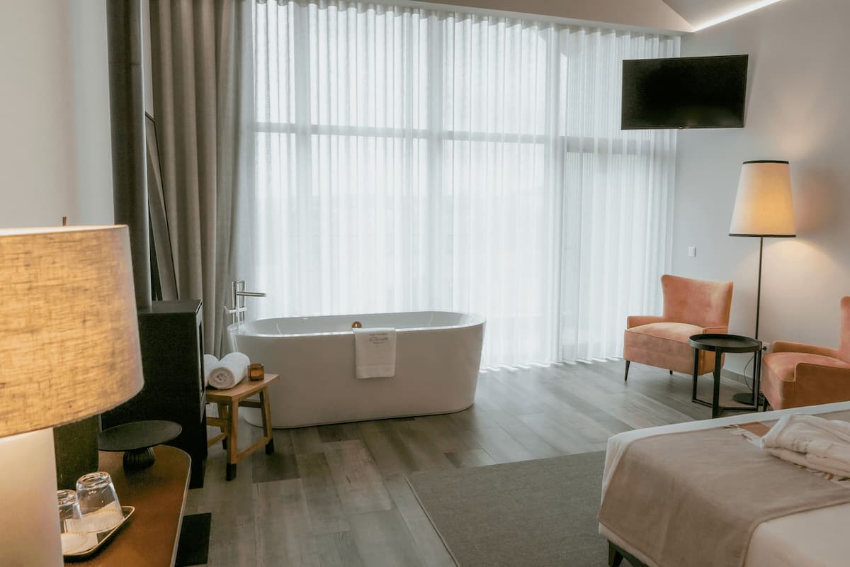 Ein modernes Hotelzimmer mit großem Fenster, einer Badewanne, einem Sessel, einem Nachttisch und einem Bett.