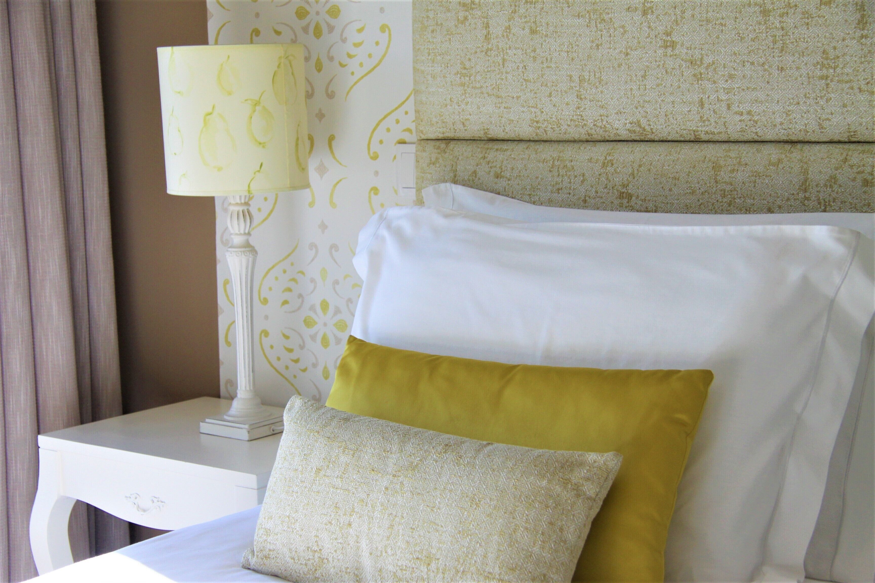 Una cama bien tendida con almohadas blancas y amarillas, una mesita de noche blanca con una lámpara y una pared decorada con un diseño.