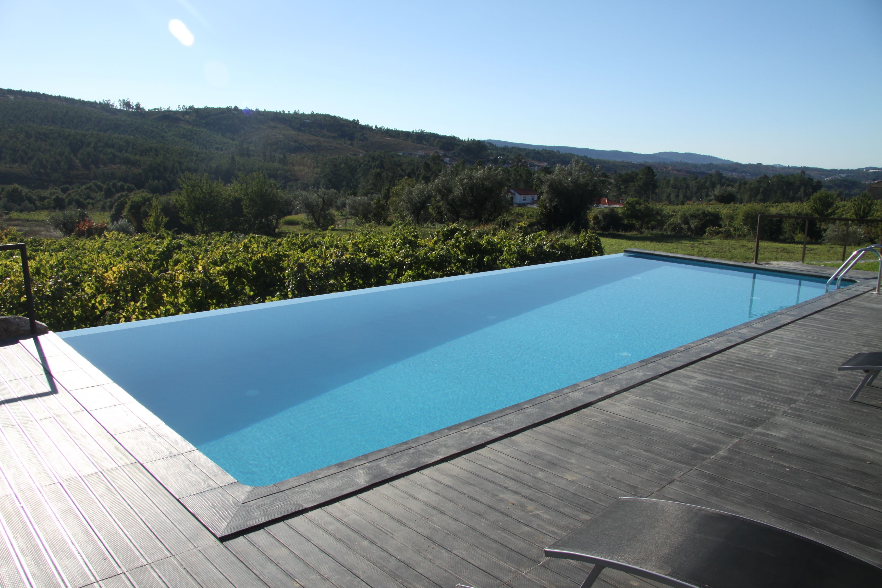 A rectangular swimming pool with a wooden deck and metal railings, surrounded by lush greenery and hills in the background.