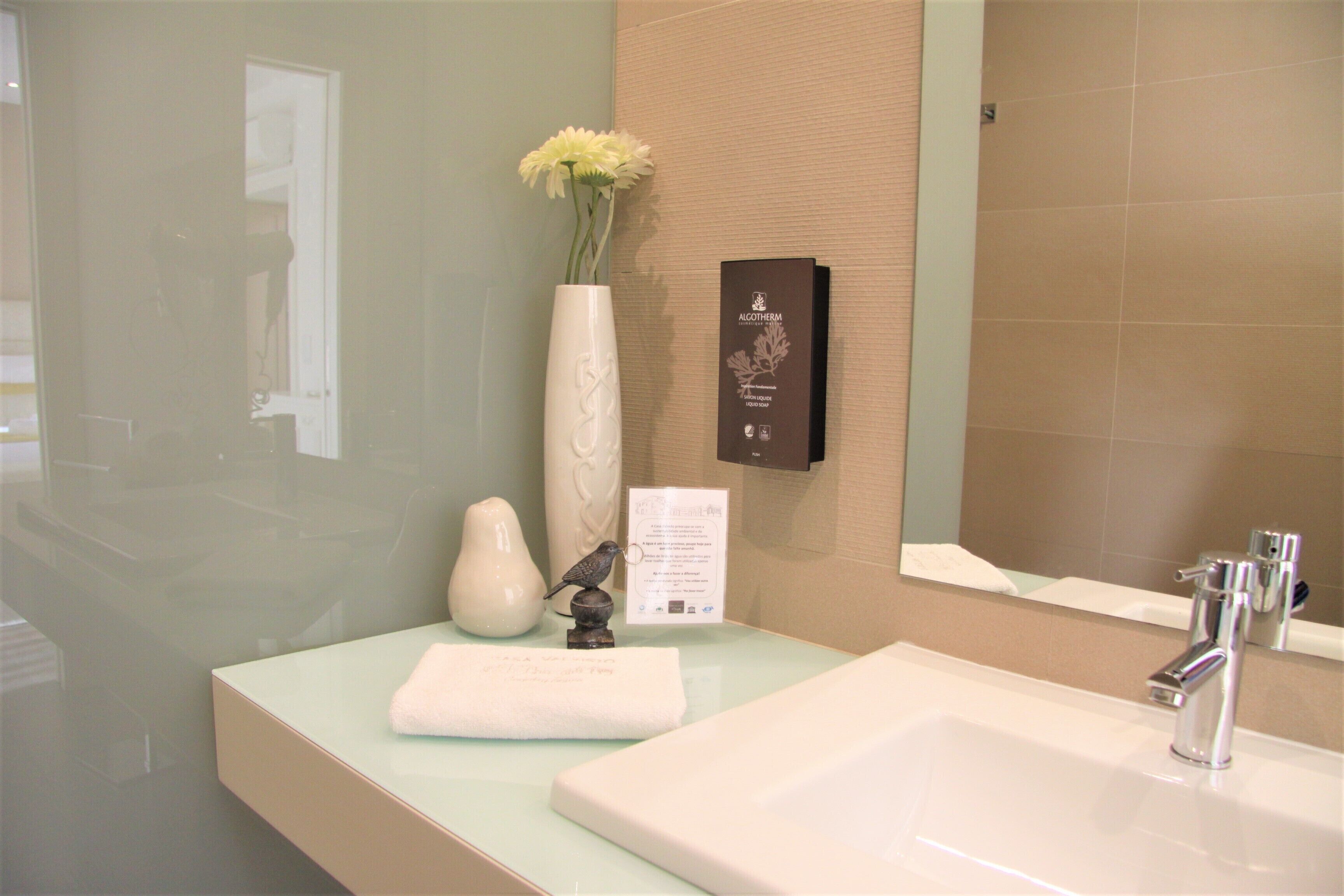 Une salle de bain avec un lavabo blanc, un miroir, un vase blanc orné de fleurs et une plaque fixée au mur.