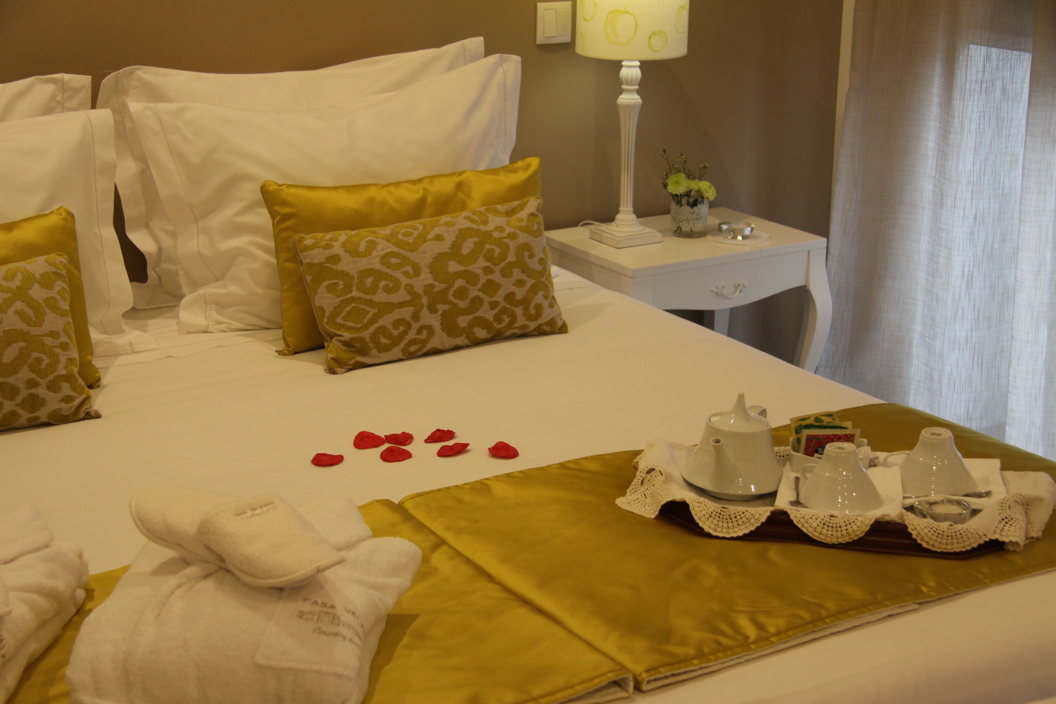 Un letto d'hotel rifatto con lenzuola bianche e dorate, un comodino con una lampada e un vaso di fiori, e un vassoio con articoli da toilette.