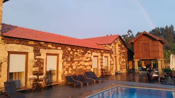 Un edificio de piedra con techo de tejas rojas, piscina y sillones de descanso.
