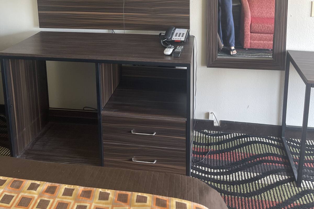 A hotel room with a dark wood desk, a flat-screen TV mounted on the wall, a telephone, and a mirror reflecting part of the room.