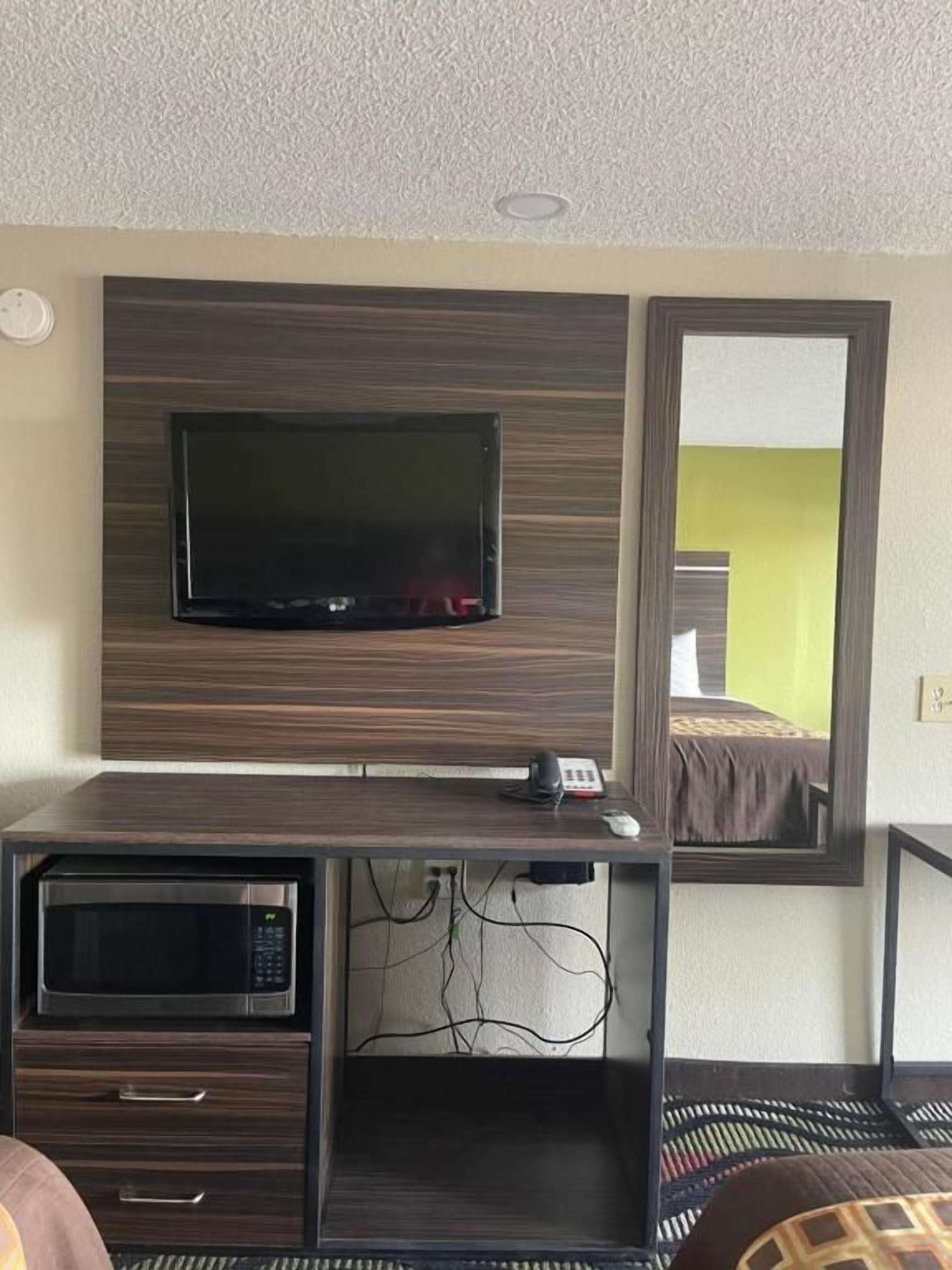Une chambre d’hôtel dotée d’un téléviseur à écran plat fixé sur un panneau mural en bois foncé, d’un bureau avec un micro-ondes et d’un lit, avec un mur vert visible dans le reflet du miroir.