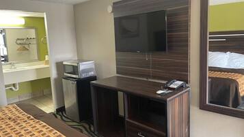 A hotel room with a bed, a desk with a flat-screen TV, a minibar, and a mirror.