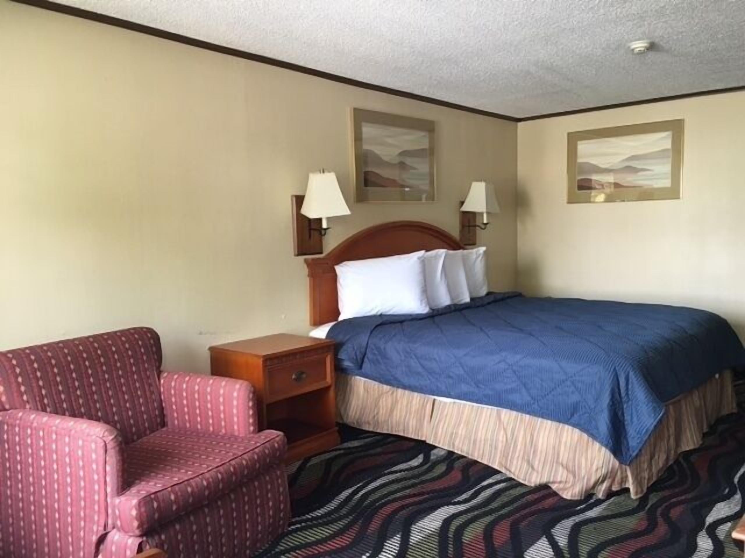 A hotel room with a bed, a chair, a nightstand, and two wall-mounted lamps.