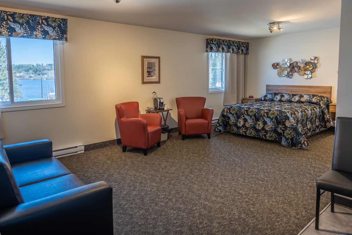 A hotel room with a bed, two red chairs, a small table with a kettle, a framed picture on the wall, and a view of the outdoors through the windows.