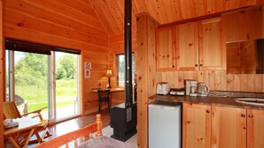 A wooden cabin with a kitchen, a dining area with a table and chairs, and a large window with a view of greenery. - Ermitage Saint-Antoine (Lac-Bouchette)