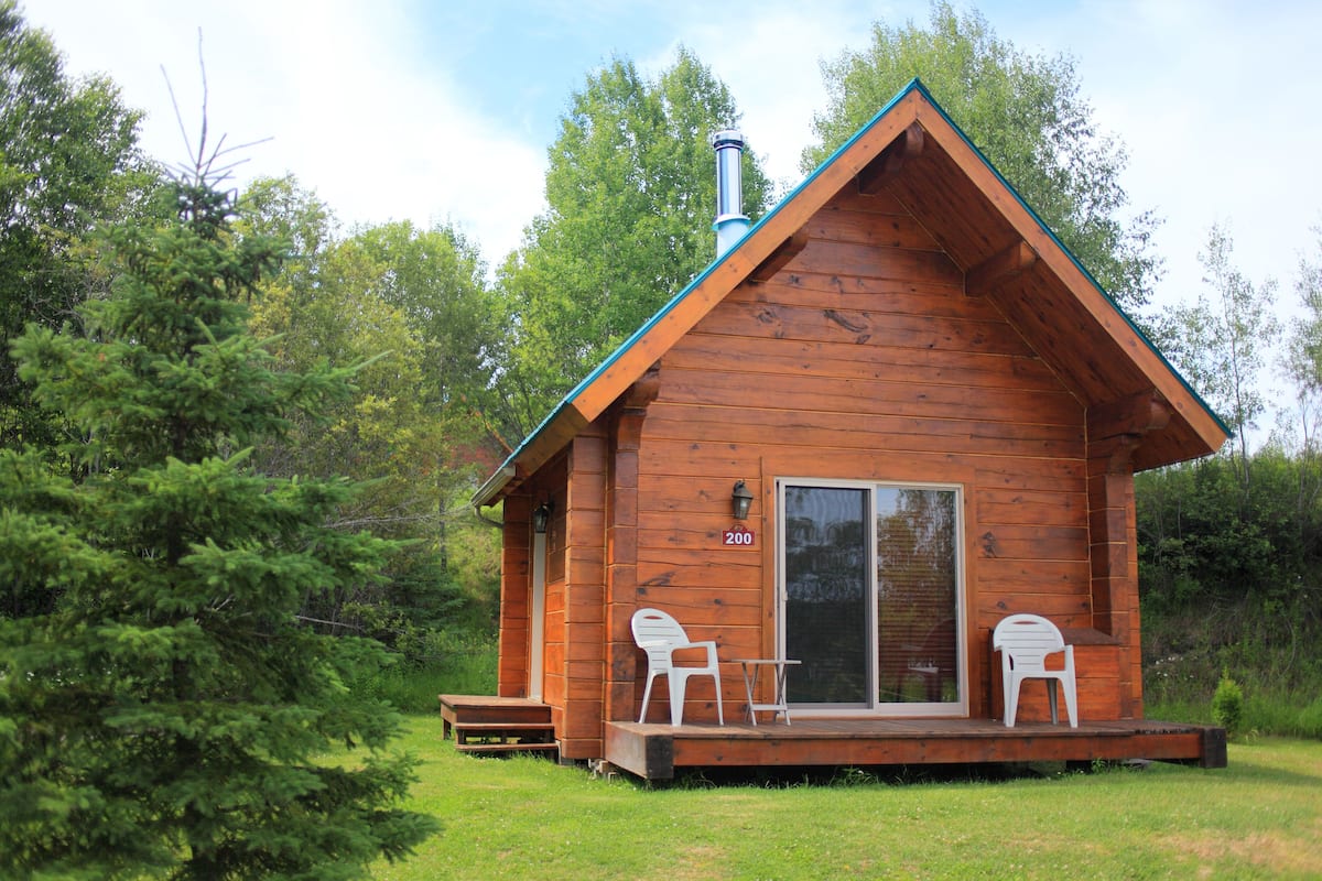 A wooden cabin with a number '200' on the door, a chimney, and two white plastic chairs on the porch.