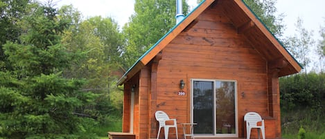 A wooden cabin with a number '200' on the door, a chimney, and two white plastic chairs on the porch.