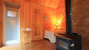 A cozy cabin room with a wooden interior, a bed, a small table with a towel, and a wood-burning stove. - Ermitage Saint-Antoine (Lac-Bouchette)