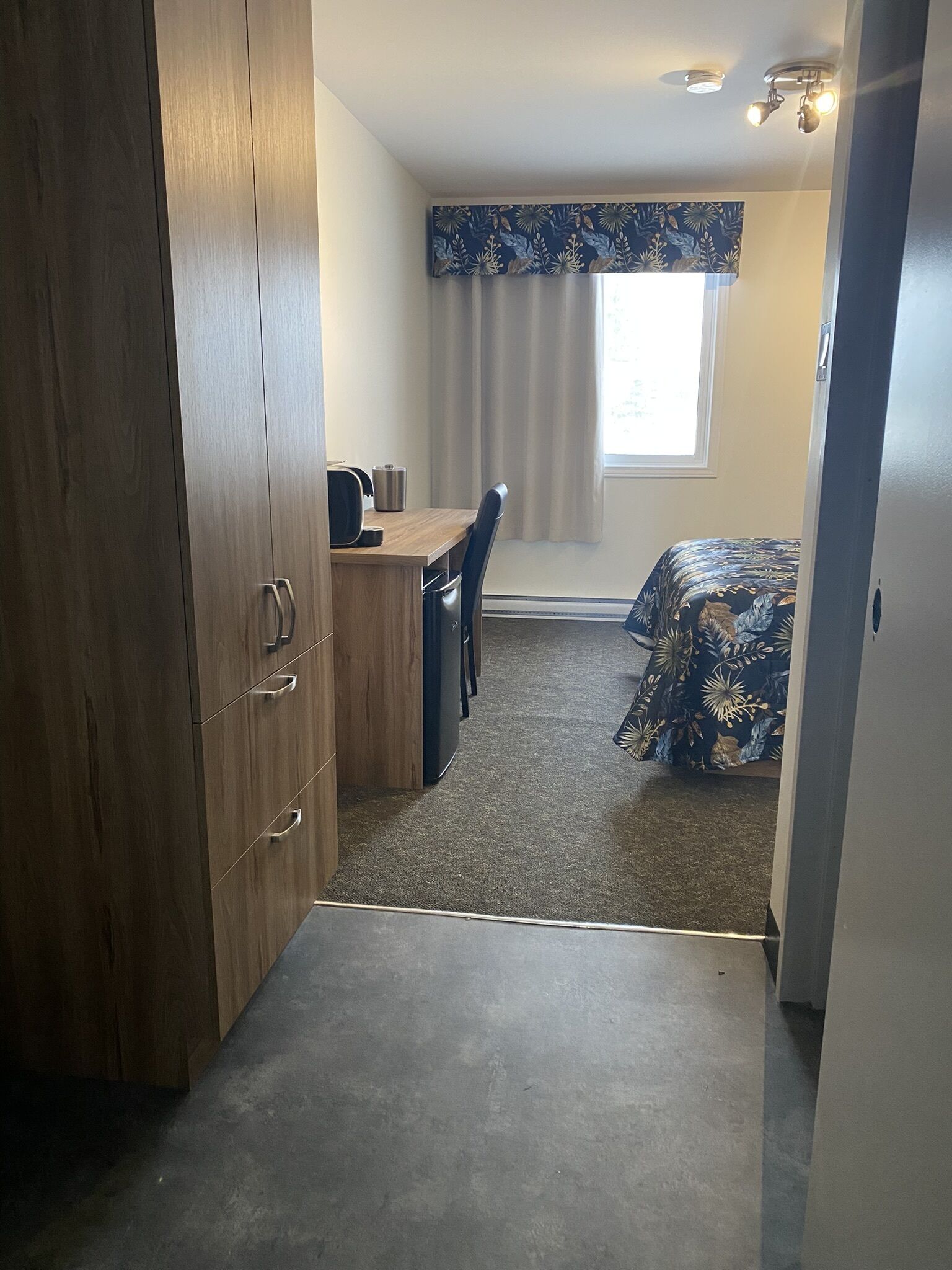 A compact room with a wooden wardrobe, a desk with a chair, a bed with a floral bedspread, and a window with curtains.