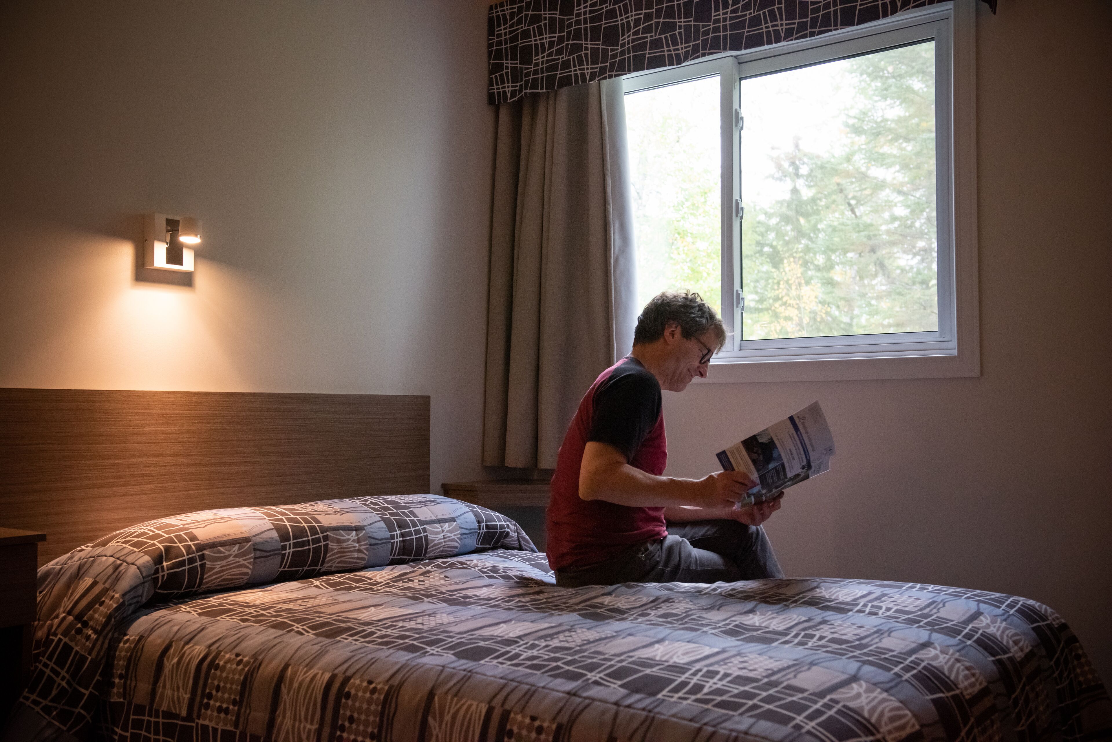 Una persona seduta su un letto in una stanza con una finestra, una lampada fissata al muro e una coperta a motivi.
