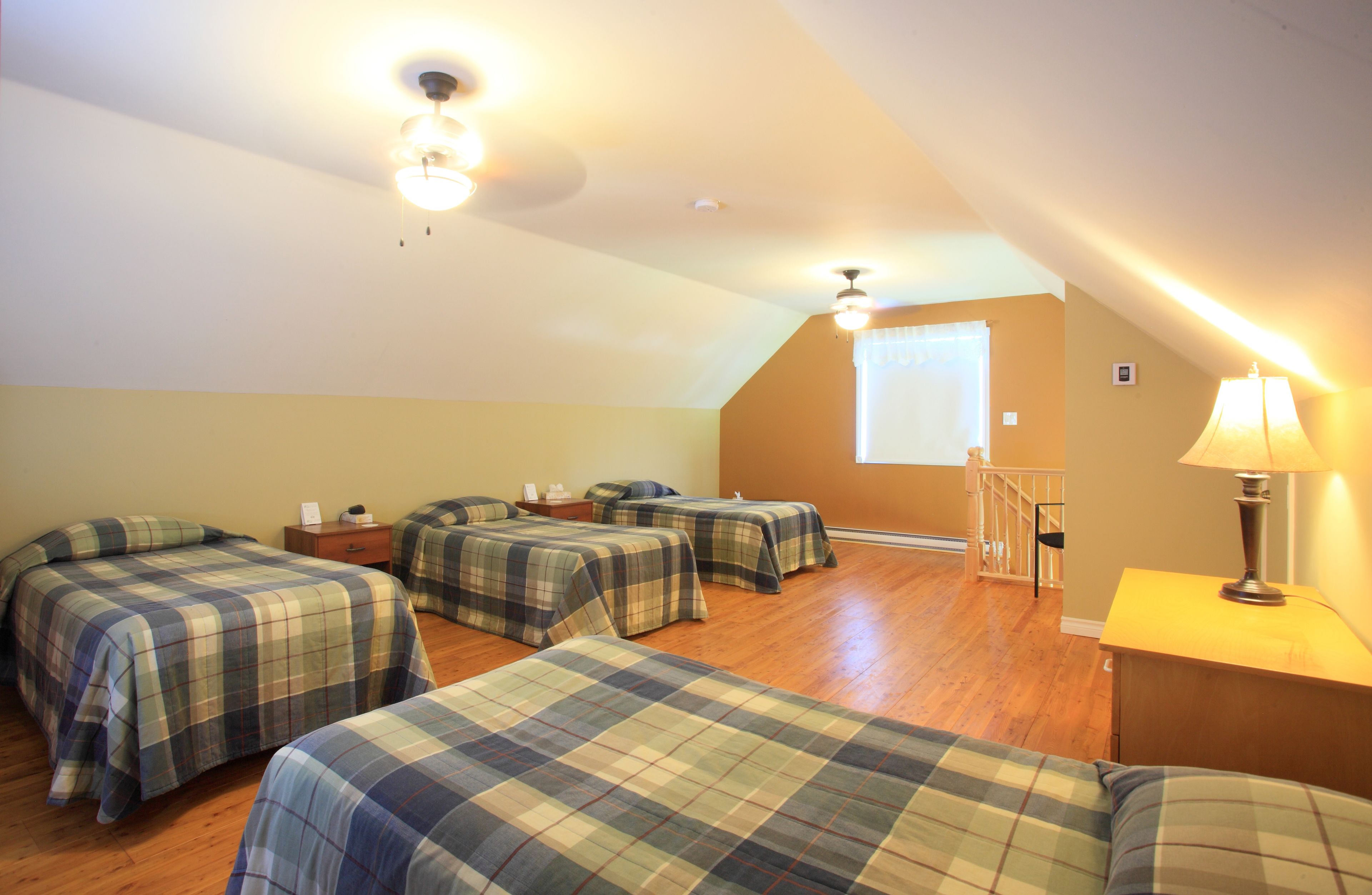 A room with four beds, wooden flooring, a ceiling fan, a window with curtains, and a lamp on a bedside table.