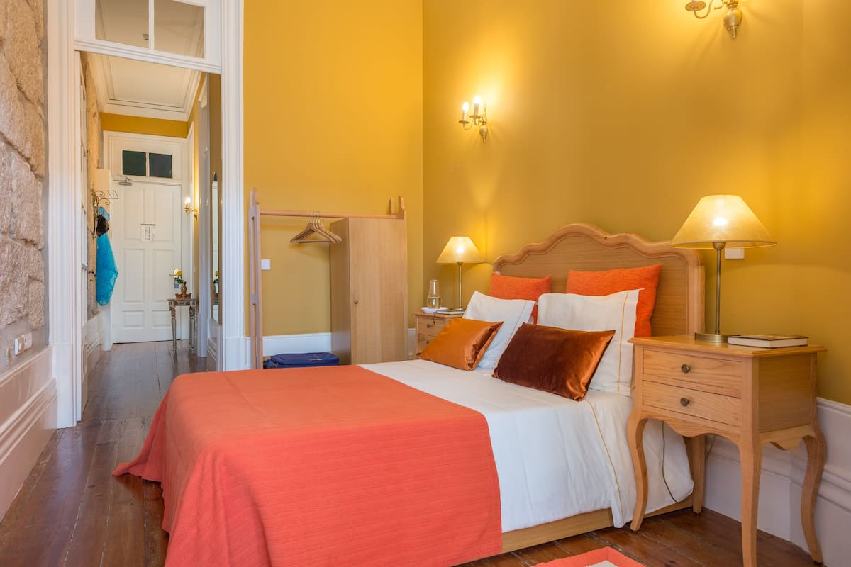 A bedroom with a bed, two bedside tables with lamps, a wooden nightstand, and a door leading to another room.
