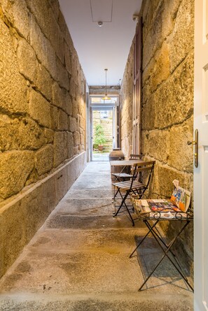 A narrow stone corridor with a table and chairs, a small display of postcards, and a view into an outdoor area with a door and a hanging light fixture.