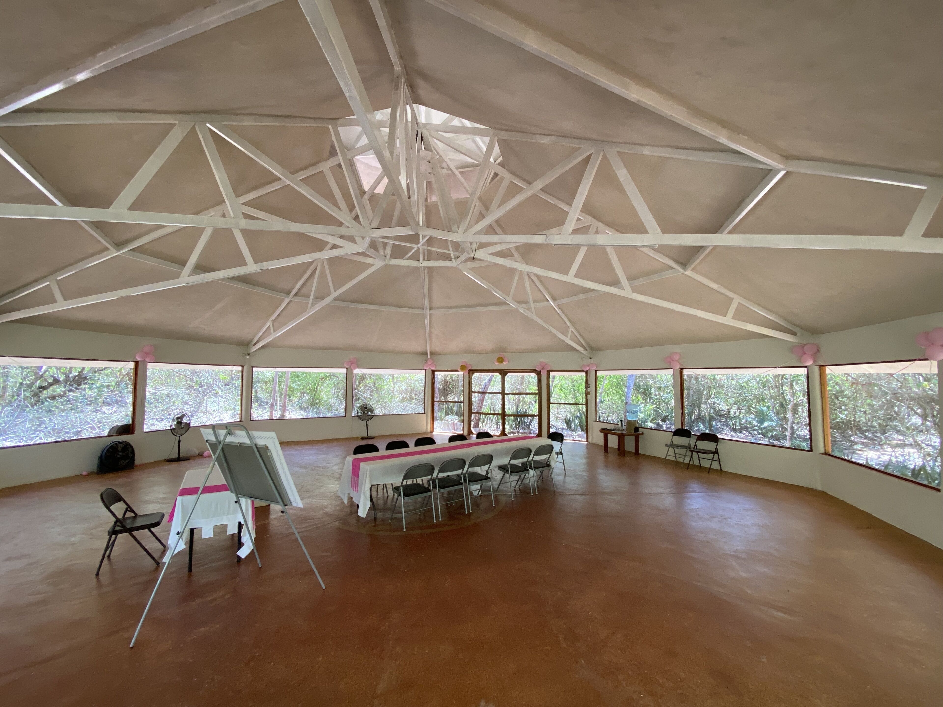 A spacious indoor area with a high ceiling and large windows. There are tables and chairs arranged for seating, and a fan is visible near the windows.
