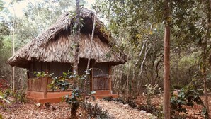 Uma cabana com telhado de palha e varanda de madeira, rodeada por uma área arborizada com pedras e plantas.