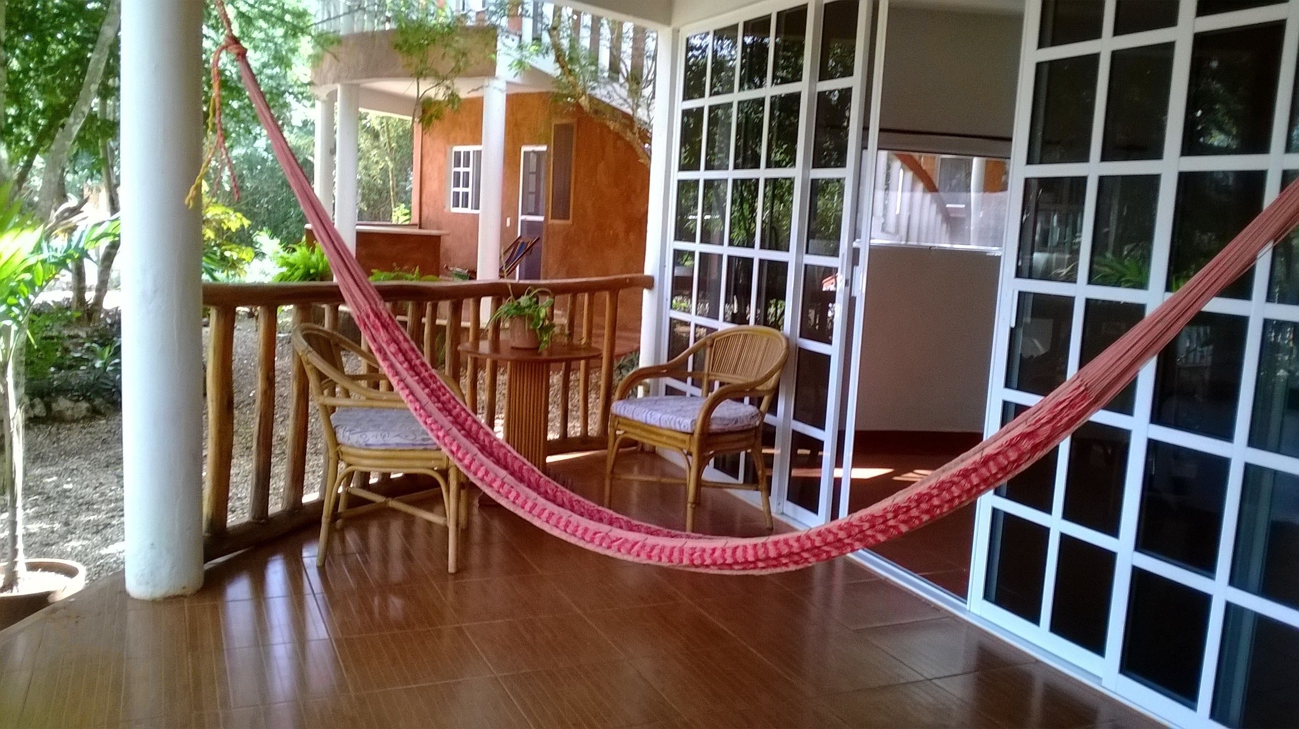 A covered patio with a wooden table and two wicker chairs, a pink hammock, and a view of a house through large windows.