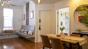 A modern living room with a wooden dining table, brown chairs, a sofa with cushions, a large window, and a brick accent wall. - 1906 Mission (San Francisco)