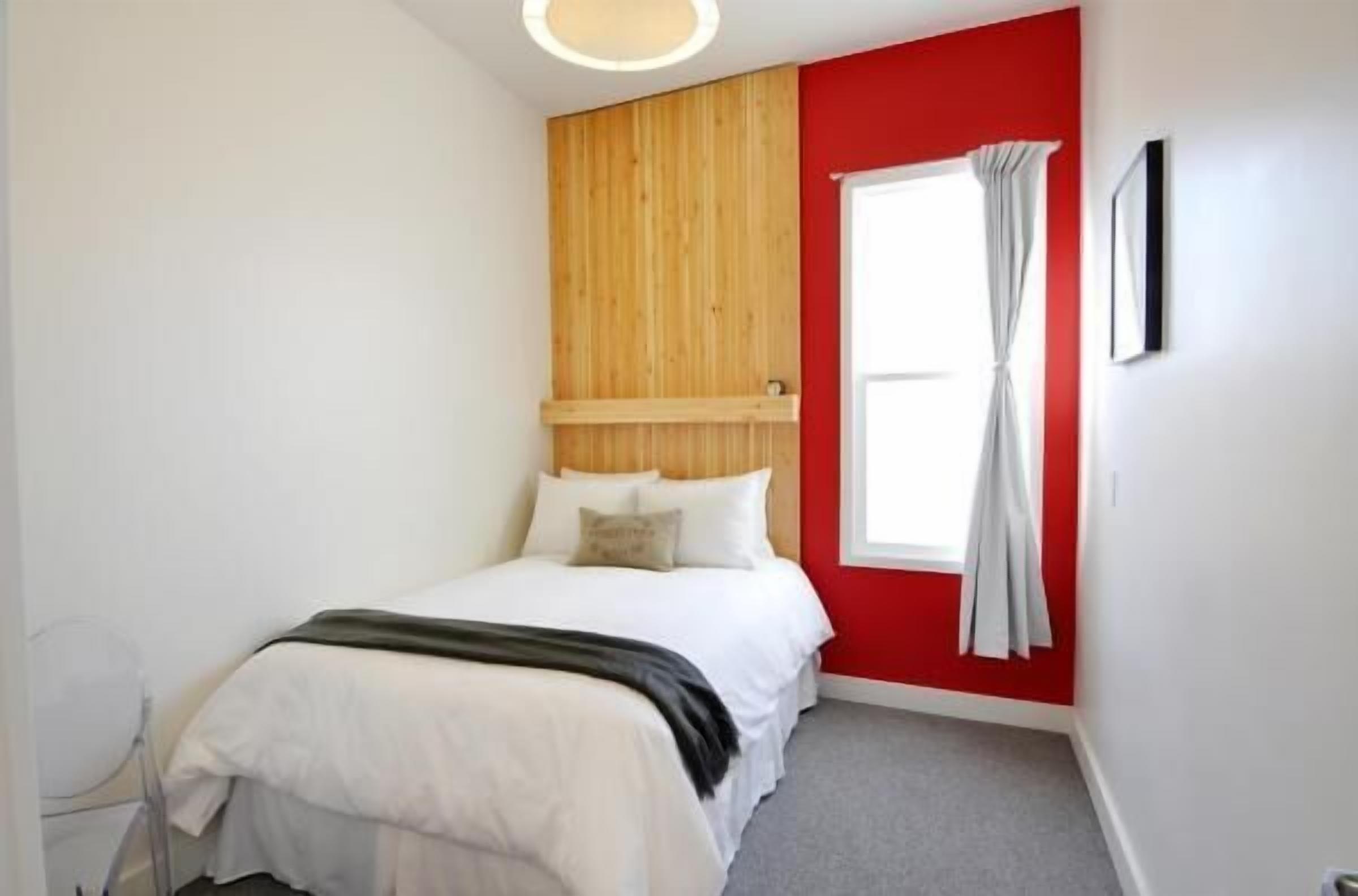 A bedroom with a bed, a wooden headboard, a window with curtains, and a red accent wall.