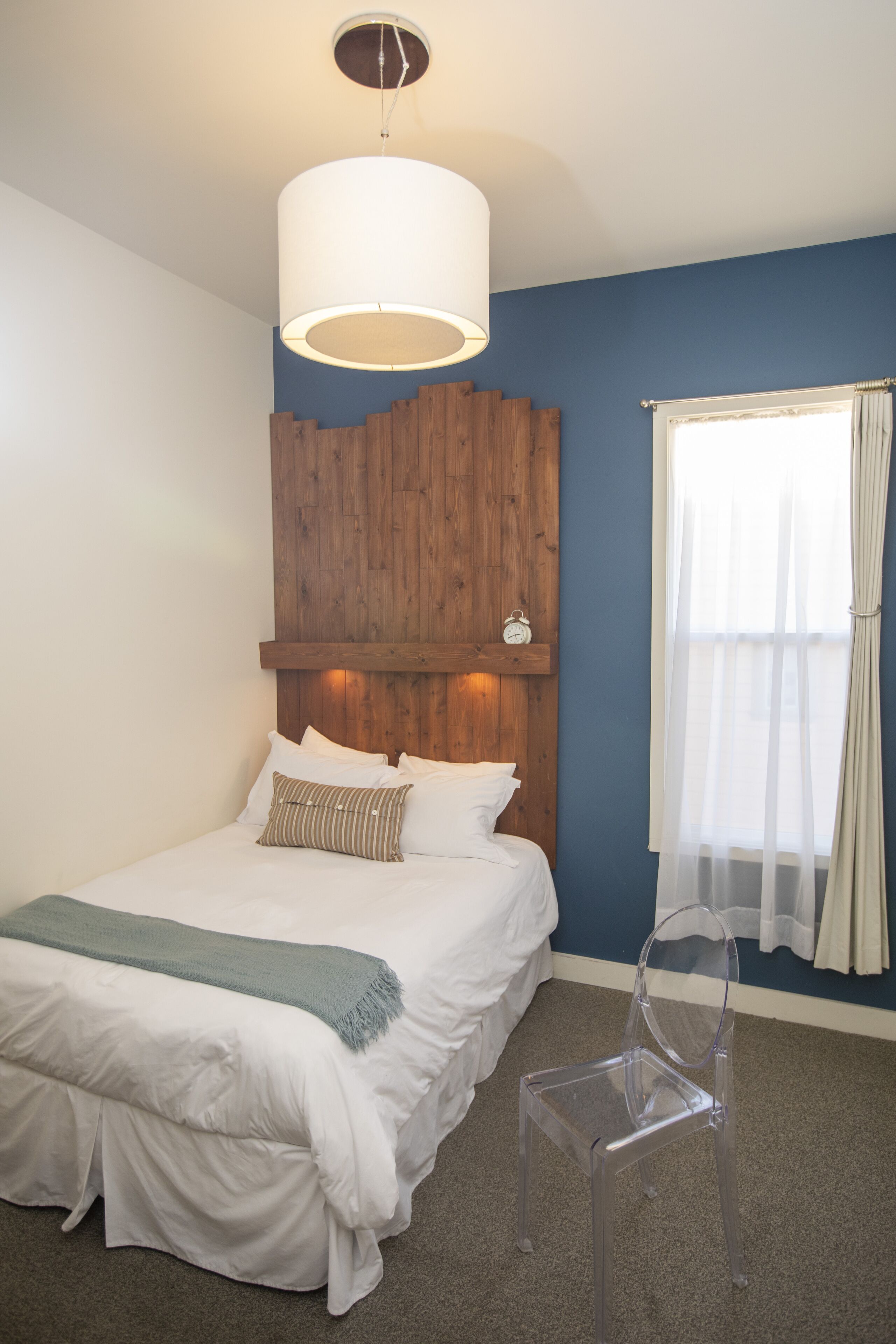 A bedroom with a wooden headboard, a bed with white bedding, a clear plastic chair, and a window with sheer curtains.