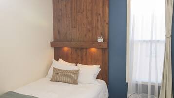 A bedroom with a wooden headboard, a bed with white bedding, a clear plastic chair, and a window with sheer curtains.