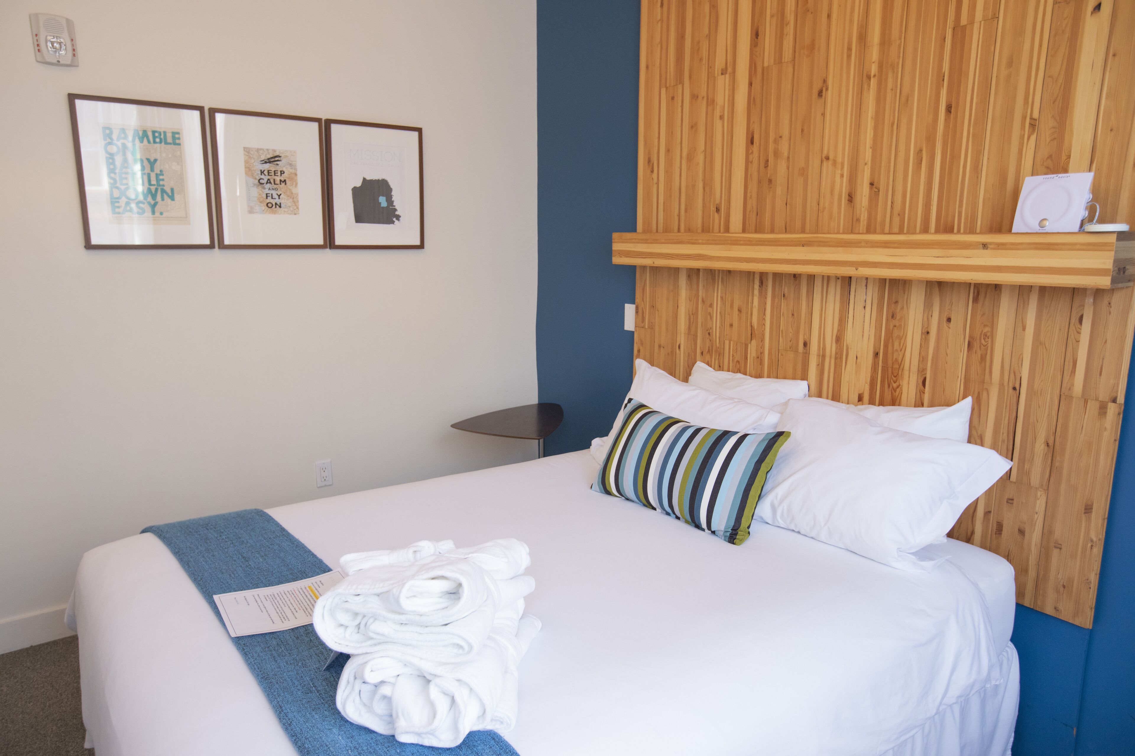A neatly made bed with white linens and a blue towel, a wooden headboard, and three framed pictures on the wall.