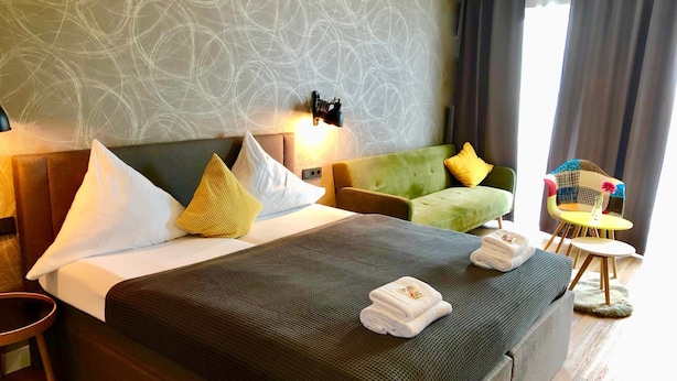 Habitación de hotel con una cama grande, un sillón verde, una mesita redonda y una pared con diseños abstractos.