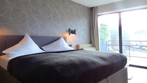 Una habitación de hotel moderna con una cama grande, un cabecero con una lámpara y vistas a la ciudad a través de amplios ventanales.