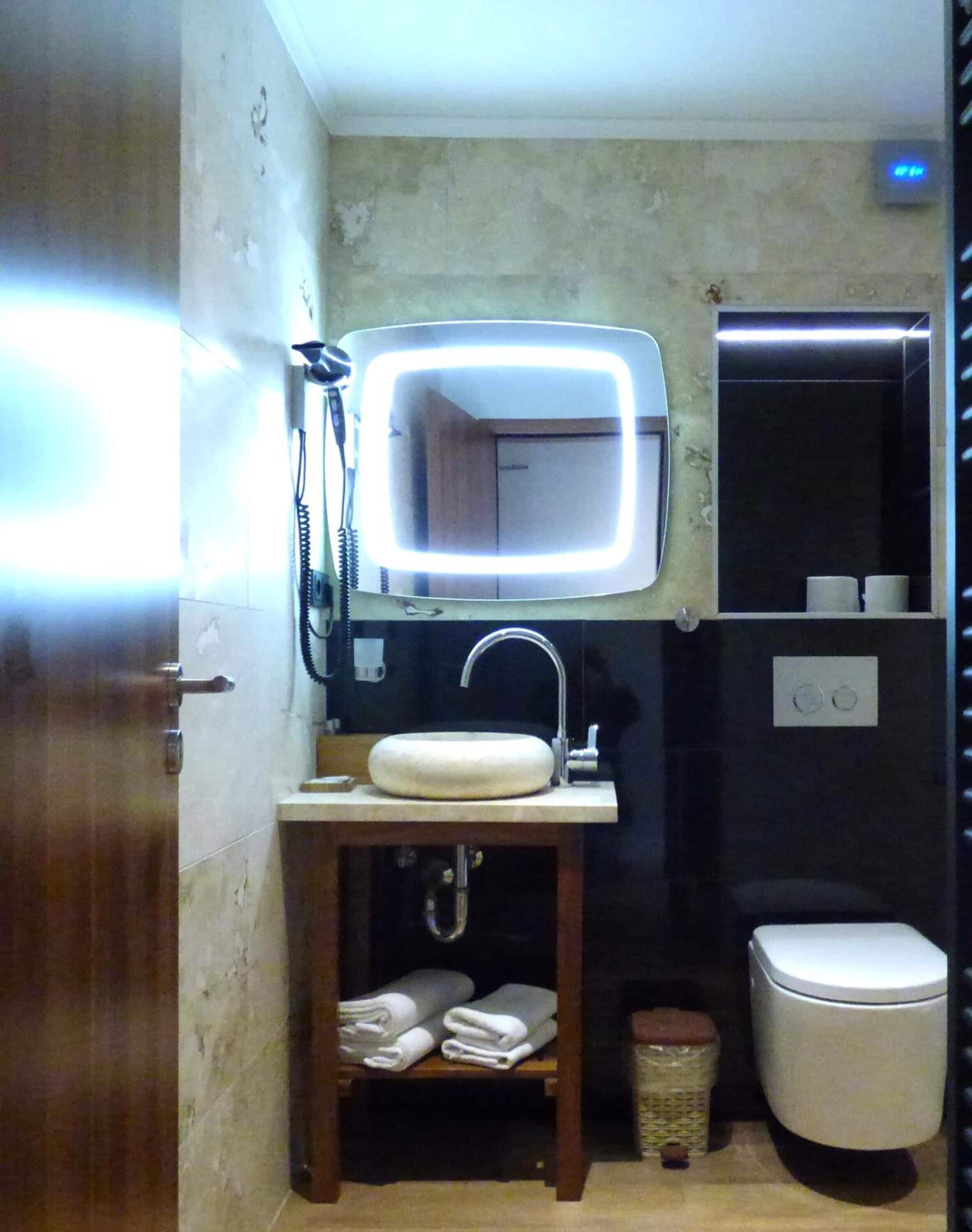 Ein kleines Badezimmer mit Waschtisch, einem Spiegel mit integrierter Beleuchtung, Toilette und Ablagefläche für Handtücher.