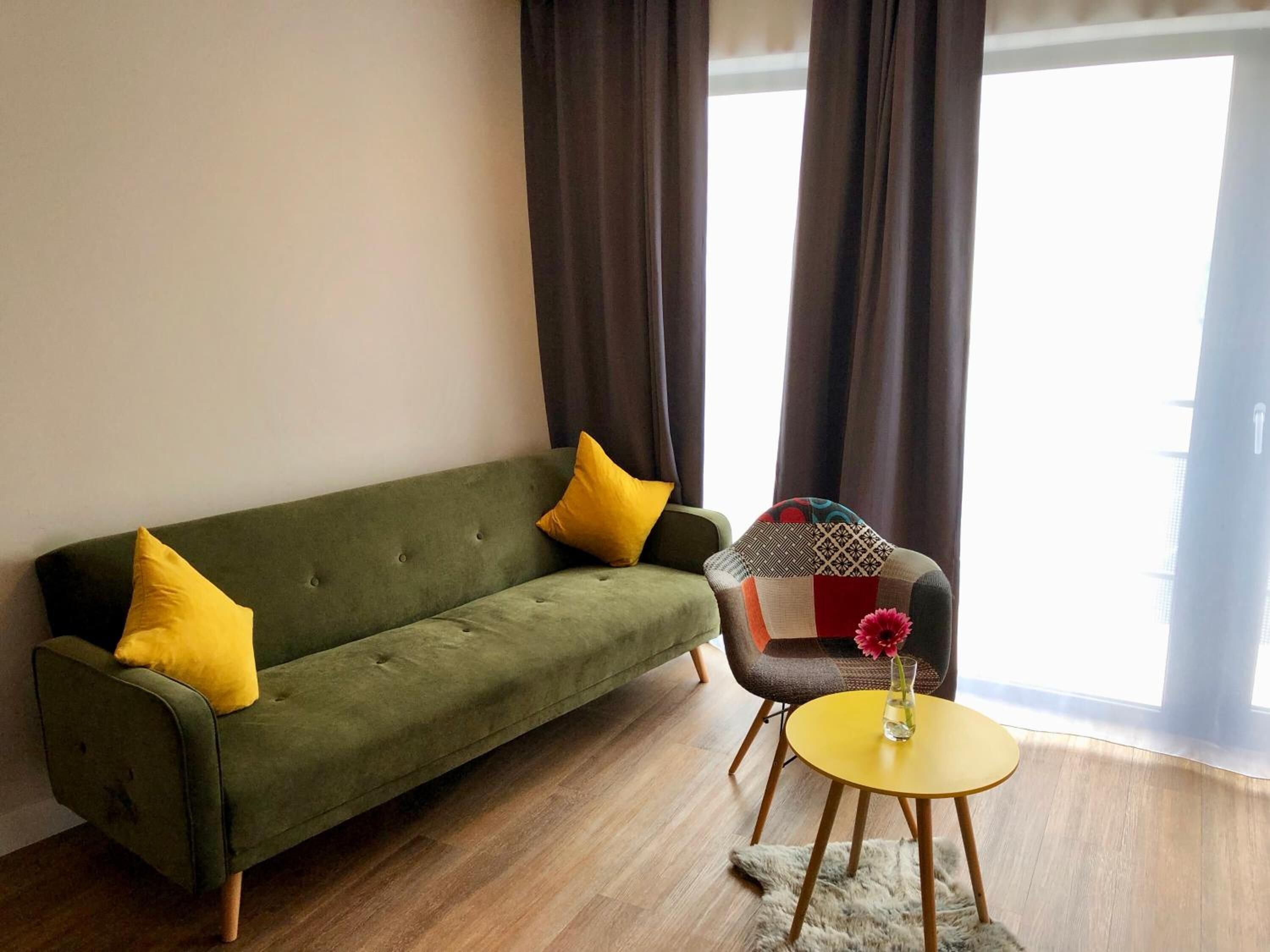 A living room with a green sofa, yellow cushions, a patterned armchair, and a small yellow table with a vase.