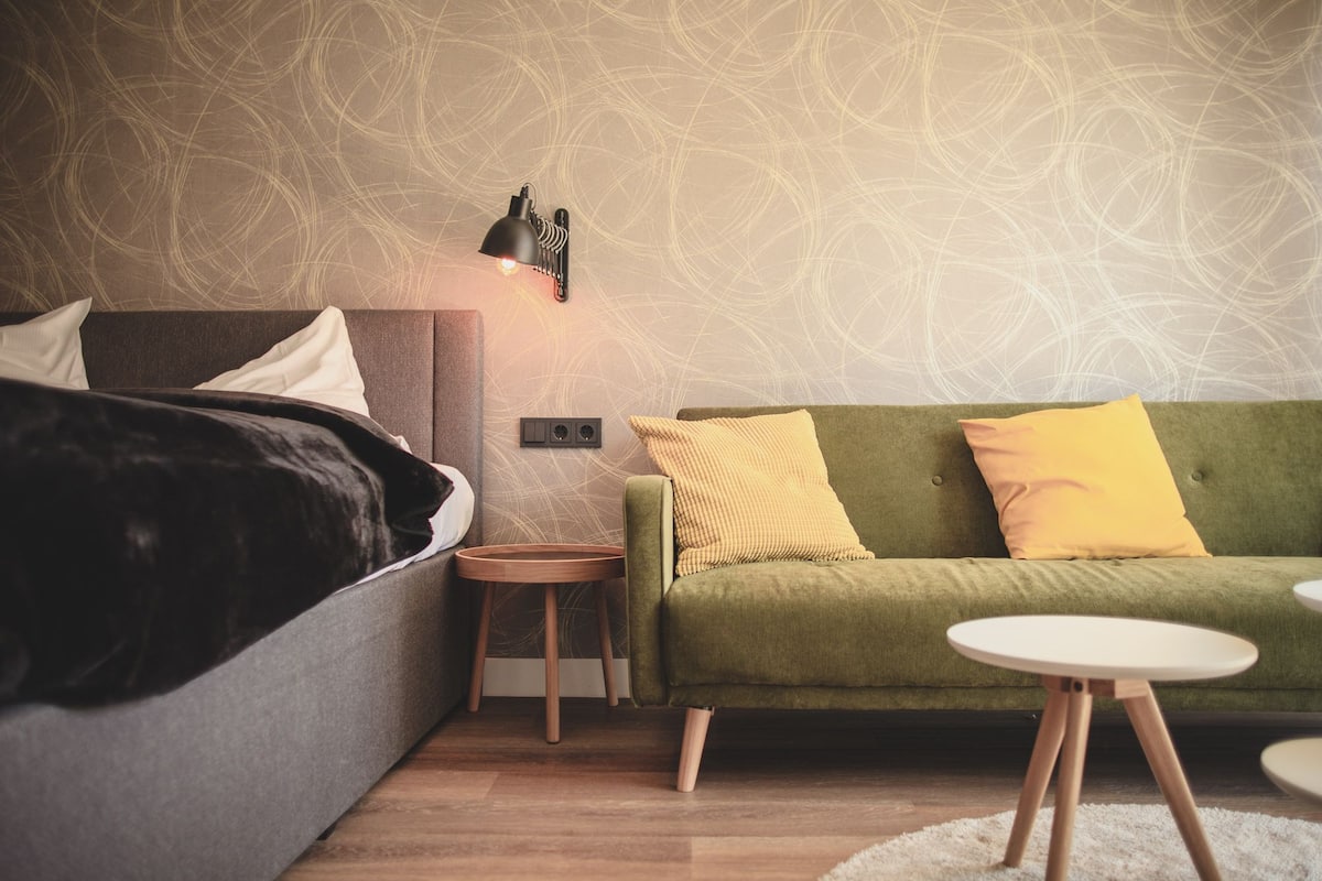 A modern bedroom with a bed, a green sofa, a small round table, and a wall-mounted light fixture.