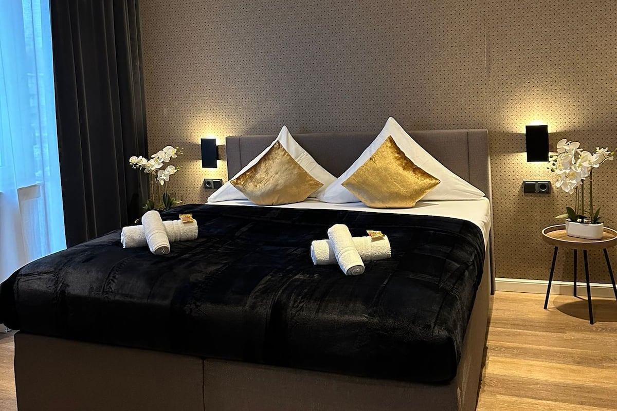 A modern bedroom with a large bed, dark curtains, and a small side table with a plant.
