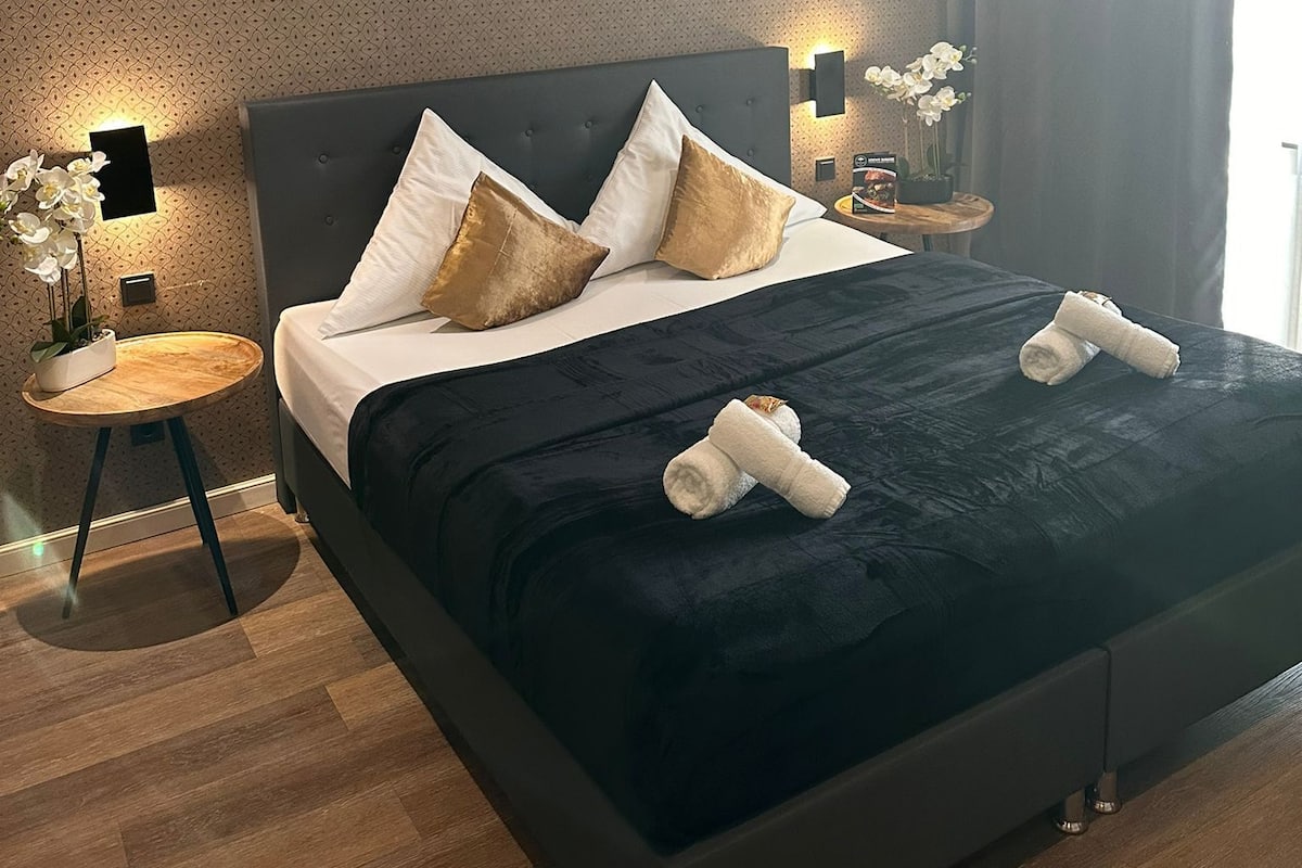 A modern hotel room with a large bed, dark bedding, and a small side table with a lamp and flowers.