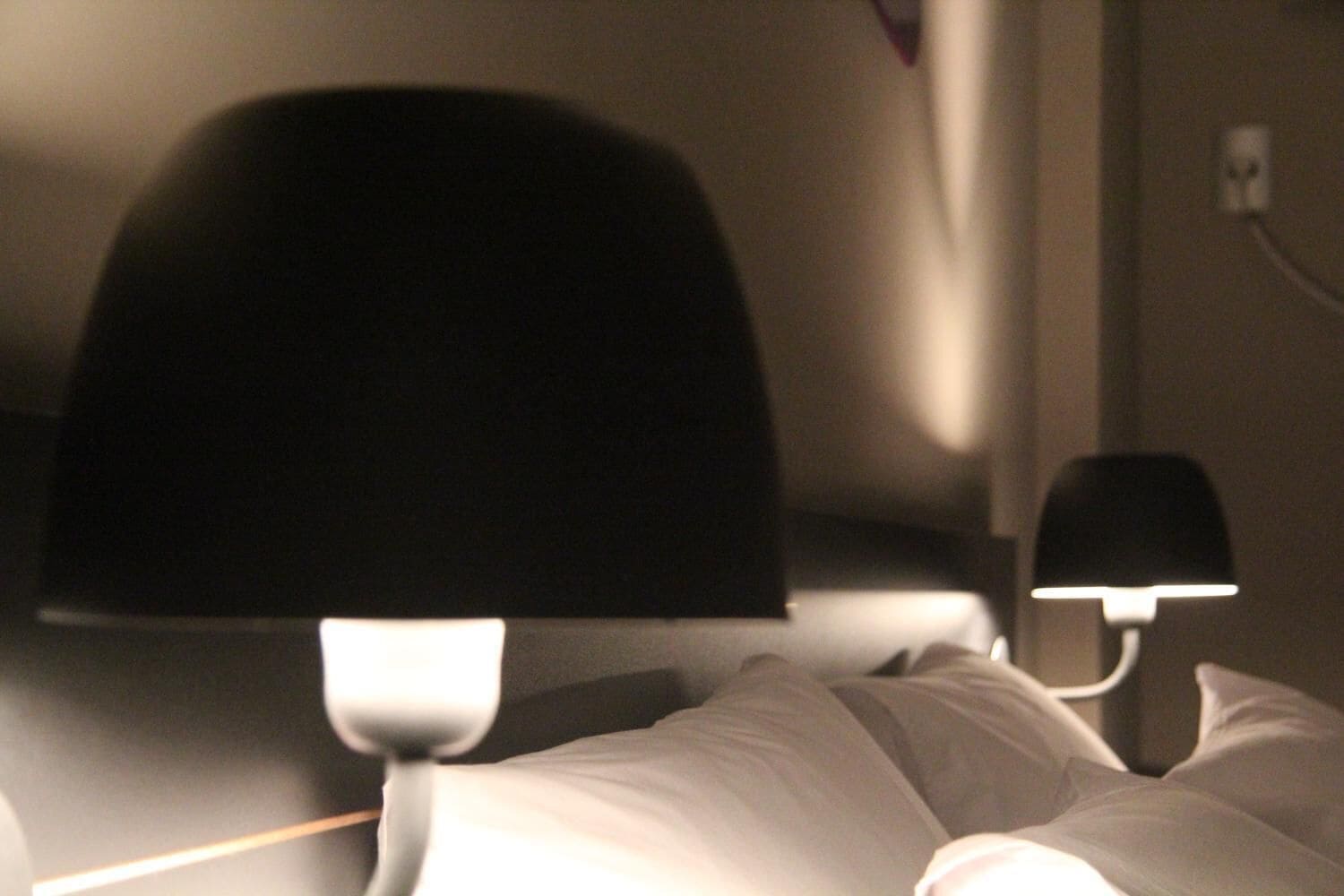 Um quarto com pouca luz, uma cama, uma mesinha de cabeceira e uma tomada na parede.