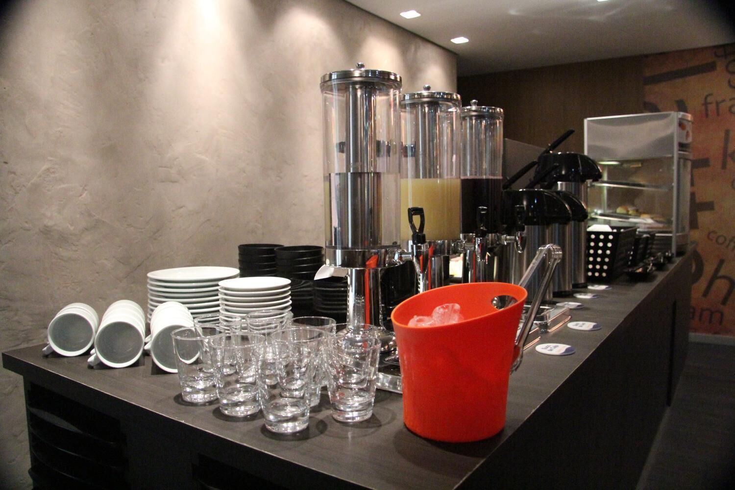 Disposição de buffet com várias estações de serviço, incluindo um balde vermelho provavelmente para gelo ou água, e diversos copos e pratos organizados para os hóspedes.