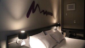 Hotellihuone, jossa on sänky, seinään asennettu ilmastointilaite ja seinätaideteos.