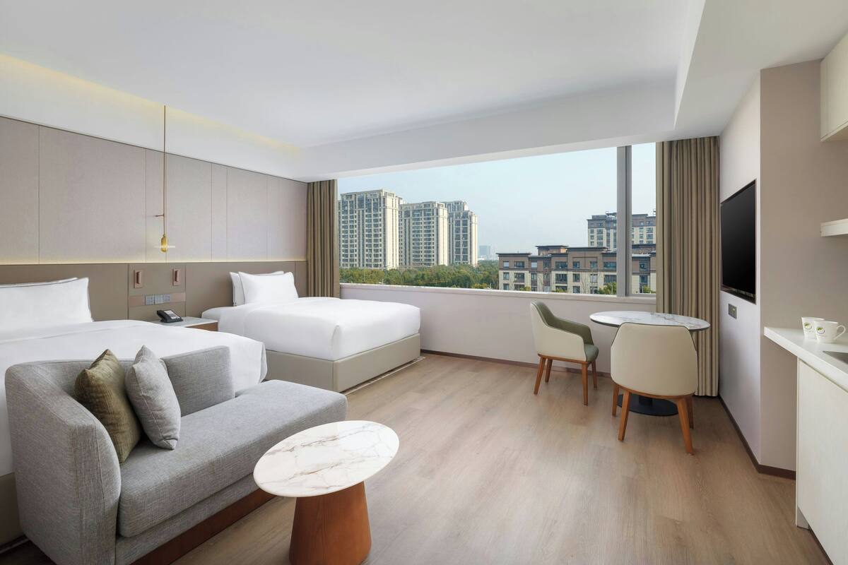 Habitación de hotel con cama, sofá, mesita, televisor y ventana con vista a la ciudad.